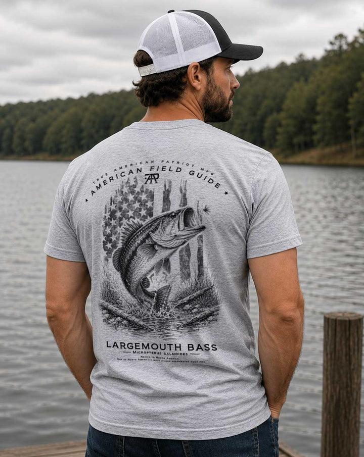 Model wearing the American Field Guide Largemouth Bass Tee in Dark Heather Gray by True American Patriot MFG. standing on a dock overlooking a lake.