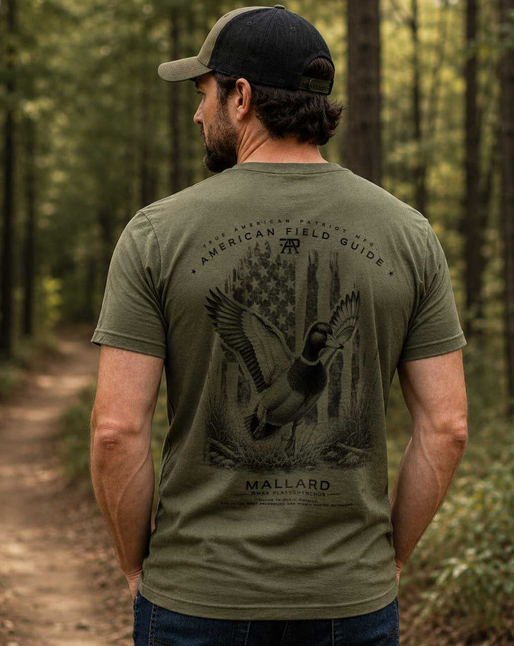 Man wearing the American Field Guide Mallard Tee in Military Green while walking through the woods, featuring a large back graphic of a mallard duck with vintage American flag artwork.