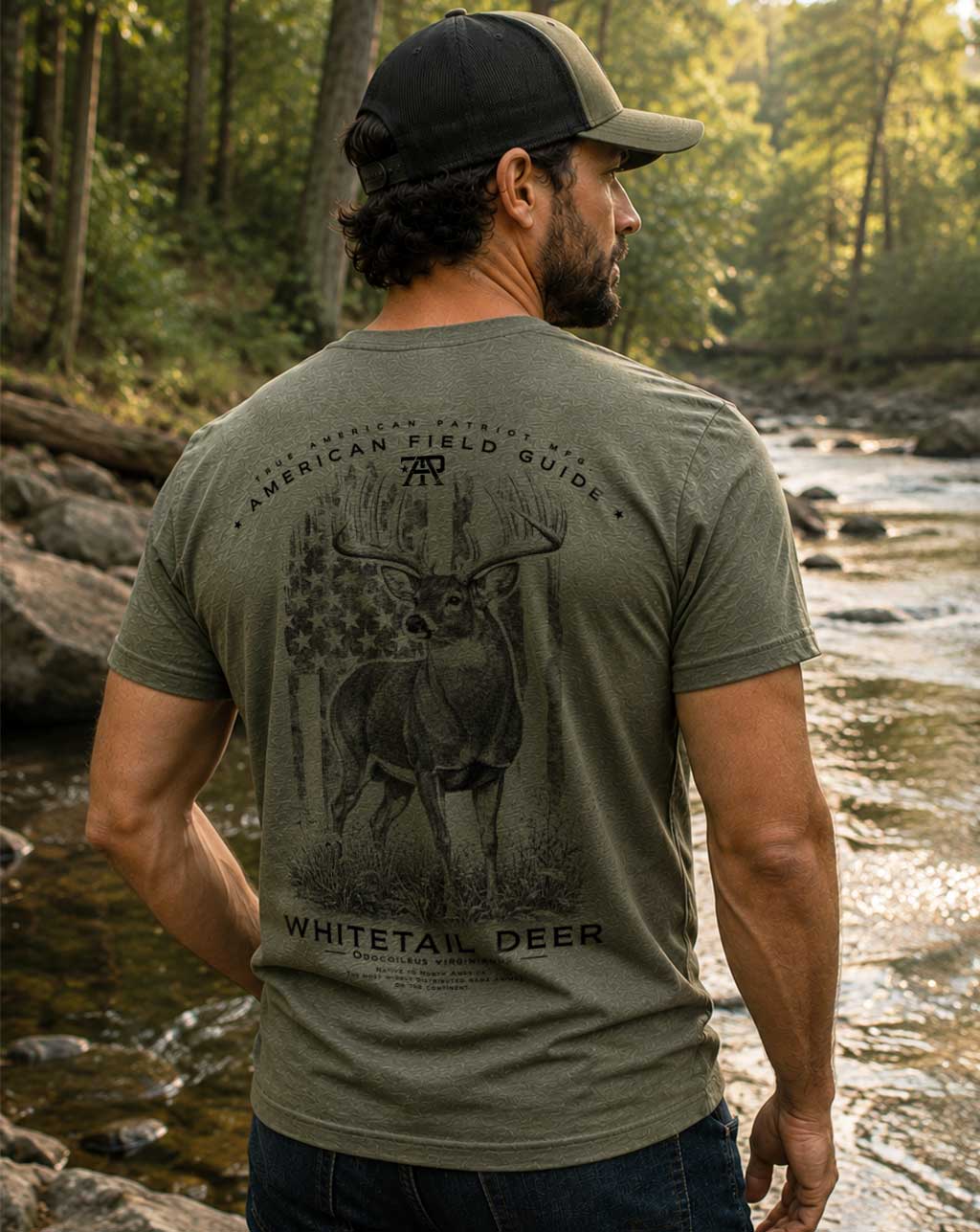 Model wearing the American Field Guide Whitetail Deer Tee in Military Green by True American Patriot MFG outdoors beside a forest stream.