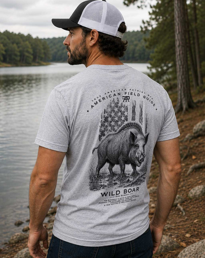Man wearing the American Field Guide Wild Boar Tee in Dark Heather Gray beside a lakeshore, featuring a large back graphic of a wild boar with vintage American flag artwork.