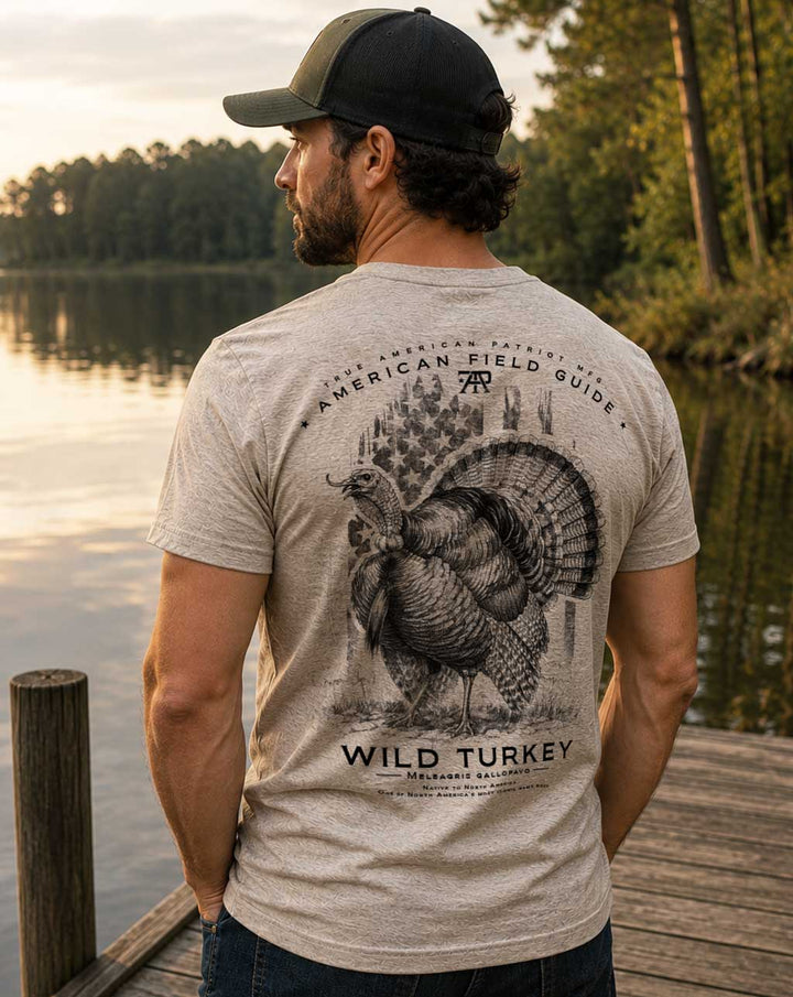 Model wearing the American Field Guide Wild Turkey Tee in Heather Tan by True American Patriot MFG outdoors beside a lake.