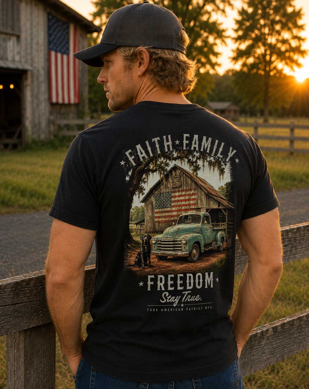 Man wearing the Faith, Family, Freedom Tee by True American Patriot MFG featuring a vintage farm truck, American flag barn, and black Labrador, photographed at sunset in a rural Georgia setting.