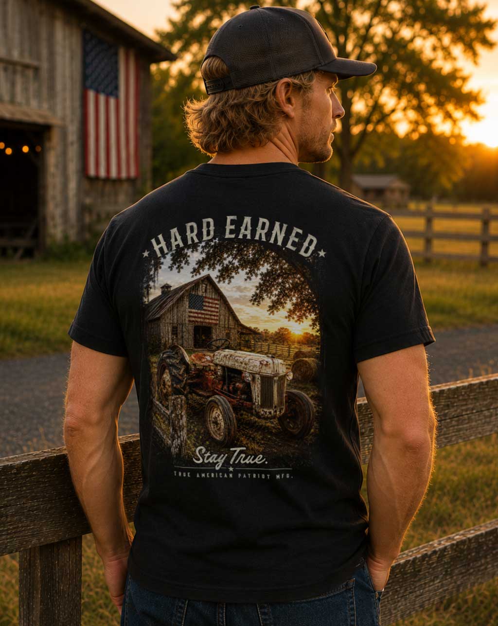 Model wearing the Hard Earned Heritage Tee featuring a vintage tractor, American flag barn, and rural farm scene. Designed and screen printed in Georgia by True American Patriot MFG.