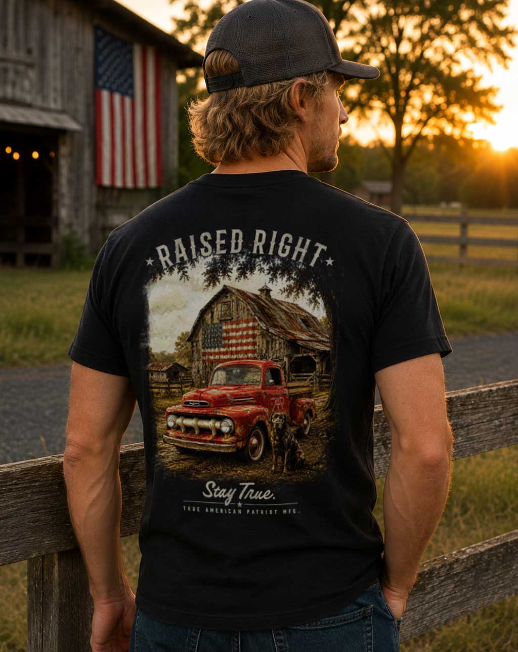 A man wearing the Raised Right Heritage graphic T-shirt featuring a vintage red pickup truck, weathered American flag barn, and hunting dog at sunset. Designed and screen printed in Georgia by True American Patriot MFG.