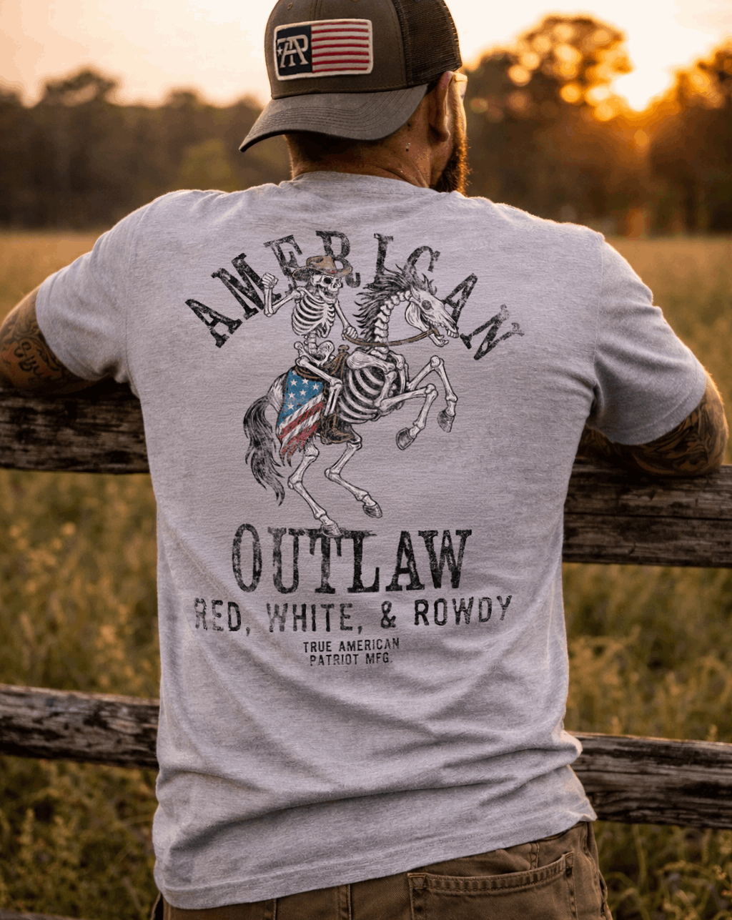 Man wearing True American Patriot Mfg Flag Patch Hat and American Outlaw - Red, White, & Rowdy Tee leaning on Fencepost watching the sunset