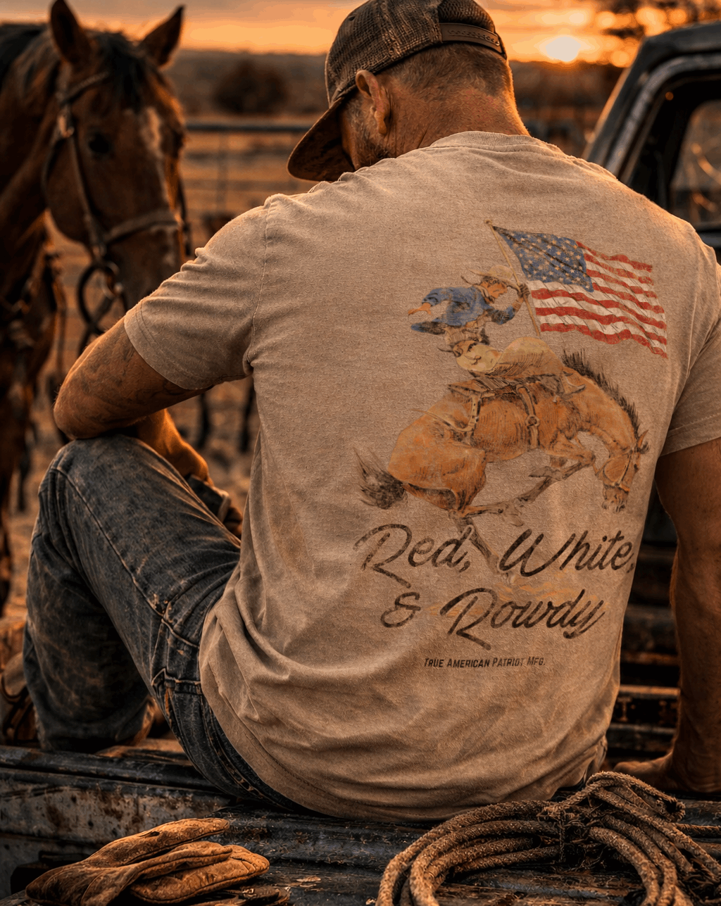 Man sitting in front of horse wearing a True American Patriot Mfg Red, White & Rowdy Rider Tee with sun setting in background