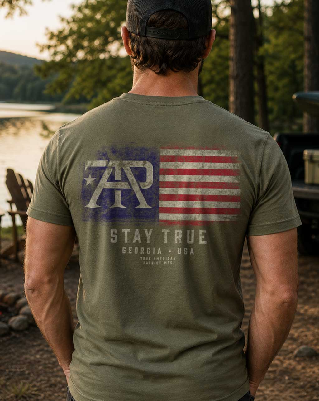 Man wearing the Stay True Flag Tee in Military Green featuring the large distressed TAP American flag back graphic, photographed outdoors by a lakeside campsite.
