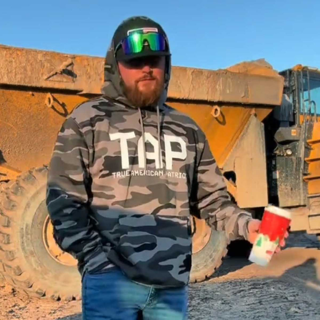 Worker wearing True American Patriot MFG camo hoodie at a construction site beside heavy equipment