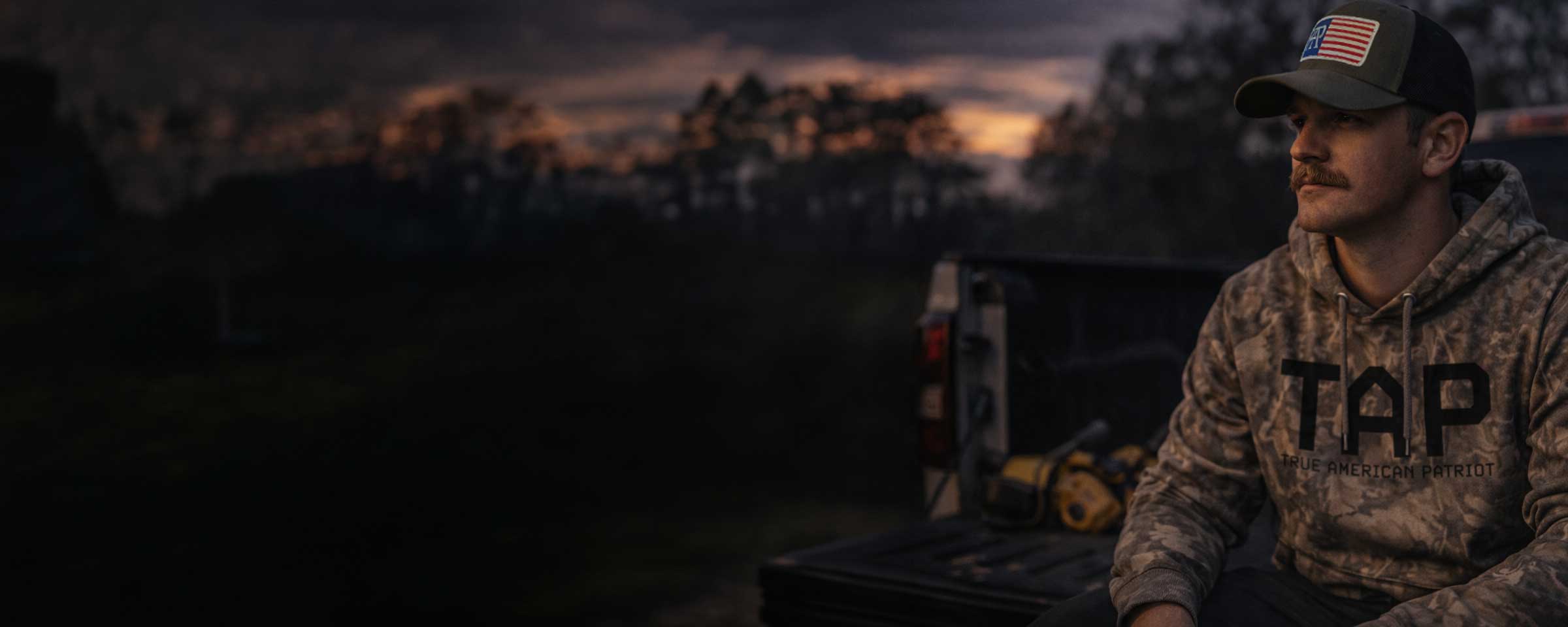 TAP camo hoodie and American flag patch hat by True American Patriot MFG in a rural outdoor setting at sunset