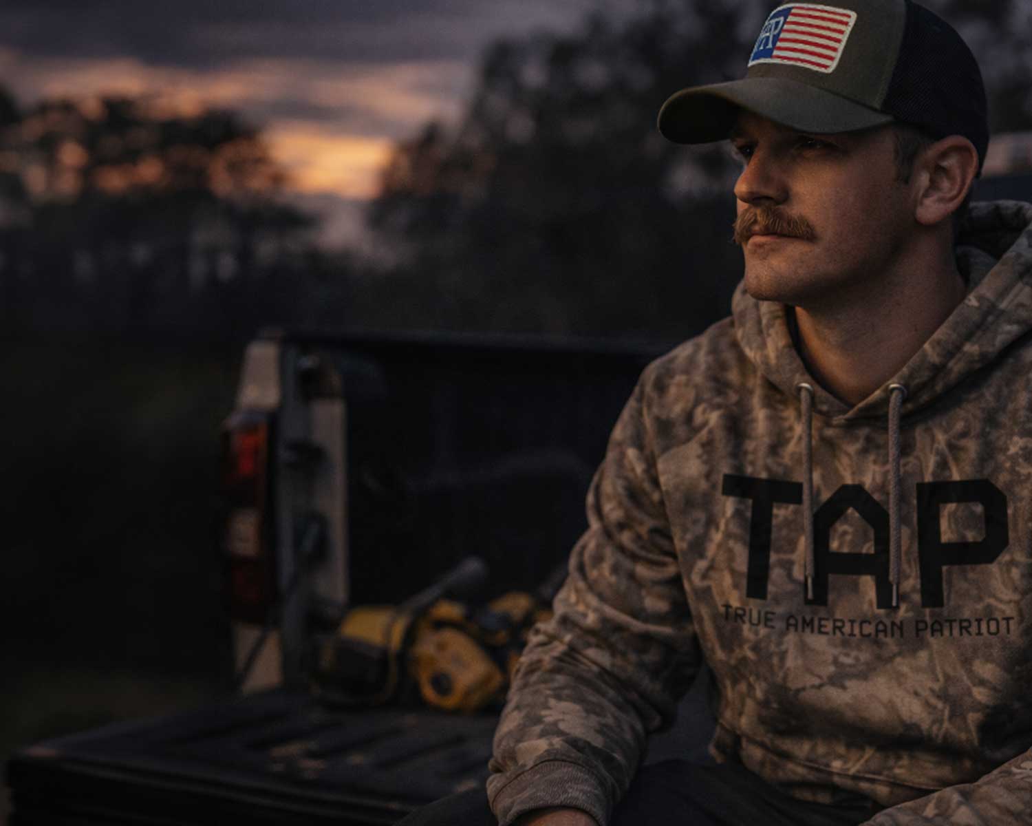 Man wearing TAP camo hoodie and American flag patch hat sitting in a pickup truck at sunset